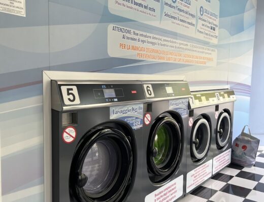 Lavanderia self-service ad Alessandria: lavatrici professionali grigie in fila con oblò nero su pavimento a scacchi, istruzioni per il lavaggio a parete. Servizio di lavaggio ad acqua.