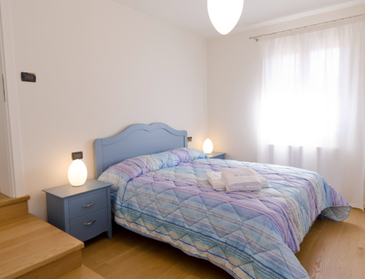 Camera matrimoniale accogliente presso Alloggi Agrituristici "Le Vignole", con letto blu, lenzuola colorate, comodini con lampade e vista luminosa dalla finestra. Ideale per un soggiorno rilassante e confortevole.