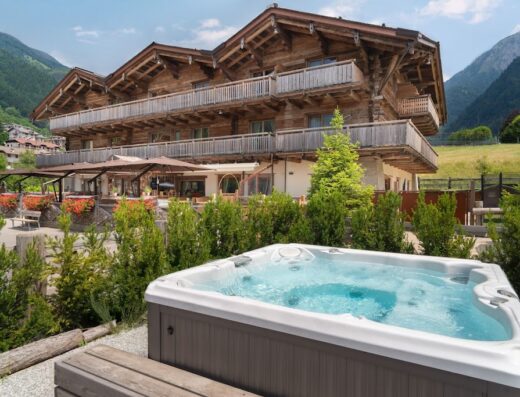 Alpen Chalet con hotel di charme e spa privata: vista di una lussuosa vasca idromassaggio con acque blu invitanti, sullo sfondo un elegante chalet di montagna in legno con balconi e ombrelloni, circondato dalla natura.
