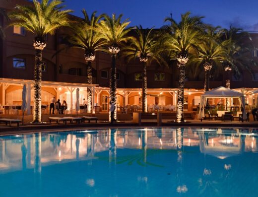Altafiumara Resort & Spa at night: Palm trees line the pool area, with illuminated buildings and lounge areas in the background, creating a serene travel destination ambiance.