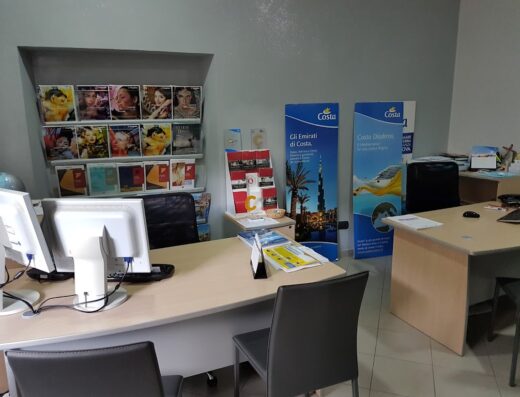 Interno dell'agenzia Alterego Viaggi di Renato Varamo, con scrivanie, computer, sedie, espositori di cataloghi di viaggi e banner pubblicitari di Costa Crociere.