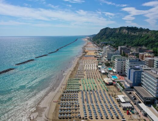 Vista aerea della spiaggia di Amadei Hotel Promenade, con ombrelloni ordinati, mare Adriatico e costa verdeggiante sullo sfondo.