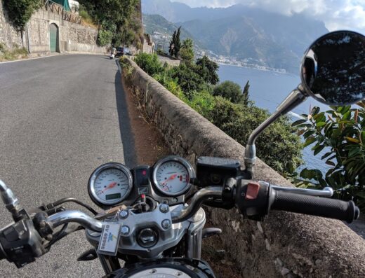Noleggio moto Amalfi: vista dal manubrio di una moto su una strada costiera con vista sul mare e sulle montagne.