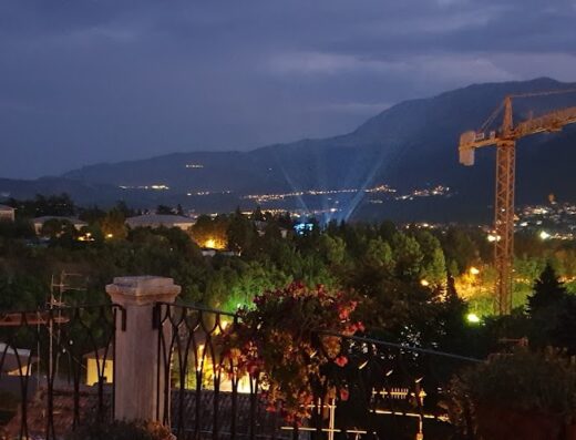 Vista notturna panoramica di una città illuminata tra le montagne, con una gru edile in primo piano e vegetazione rigogliosa.