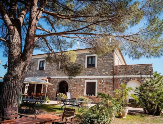 Anna dei Sapori: Esterno di un edificio in pietra con tavoli e sedie da esterno sotto un grande albero, atmosfera accogliente.