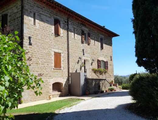 Exterior view of Antica Residenza Montereano, a stone building with brown shutters, situated in a rural setting with trees and a gravel driveway.