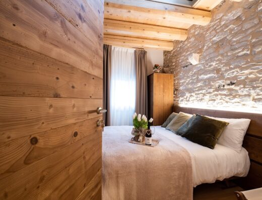 Camera matrimoniale con letto ornato da fiori e bottiglia di vino, pareti in pietra e dettagli in legno all'Antico Borgo Brunelli.