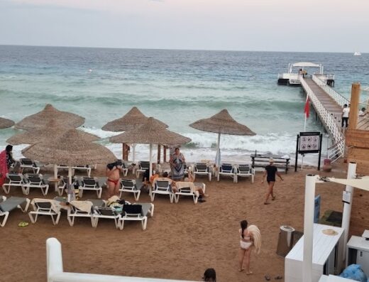 Affollata spiaggia ad Antigua con ombrelloni di paglia, lettini prendisole e bagnanti che si rilassano vicino al mare cristallino e ad un pontile attrezzato.