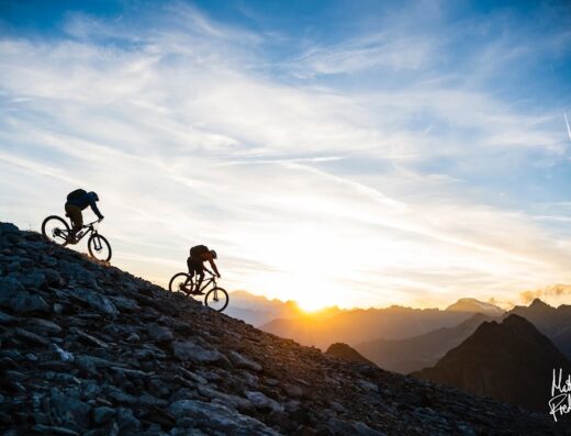 Due ciclisti in mountain bike praticano freeride su un terreno roccioso in Valle d'Aosta al tramonto, offrendo servizi e accessori per appassionati di viaggi e avventura.