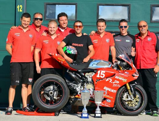 Team AP Racing Moto con moto Ducati rossa numero 15 e due trofei, meccanici e pilota sorridenti sullo sfondo di un garage.