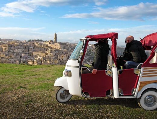 Ape Calessino motorcycle tour in Matera, Italy, with two passengers enjoying the view of the Sassi di Matera, a UNESCO World Heritage site. The unique transportation and historic landscape offer an unforgettable travel experience.
