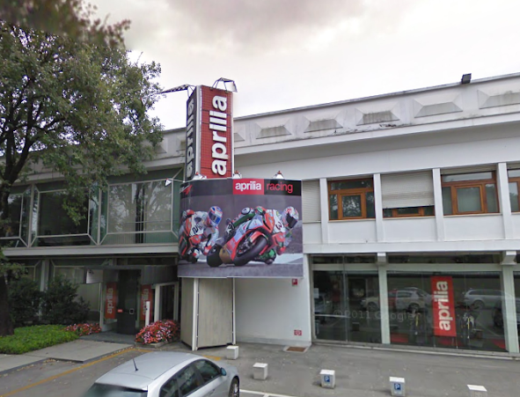 Aprilia Factory Outlet exterior with Aprilia Racing banner, motorcycles displayed in windows, and people browsing; promoting Aprilia motorcycle sales and service.