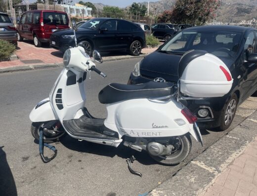 Noleggio scooter: Vespa bianca parcheggiata con bauletto e antifurto, pronta per esplorare la zona. Servizio di ar-rent scooter.