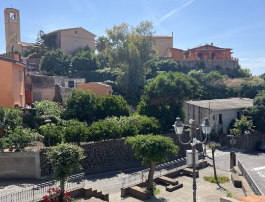 Vista panoramica di Arcu de Chelu B&B, con case colorate, una torre campanaria, vegetazione lussureggiante e dettagli architettonici tipici della Sardegna.