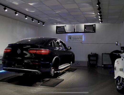 Armenise Vehicle Care Modugno Z.I.: Interno dell'officina con un'auto di lusso nera senza ruote su un ponte sollevatore e una Vespa bianca in un angolo.