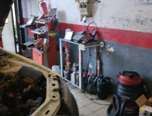 Interno di un'officina meccanica con un'auto in primo piano, attrezzature di riparazione auto e strumenti di servizio visibili.