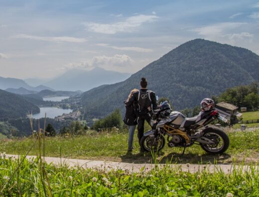 Una coppia di motociclisti ammira il panorama montano del Trentino con la loro moto Aprilia Dorsoduro, accessoriata con caschi e abbigliamento tecnico.