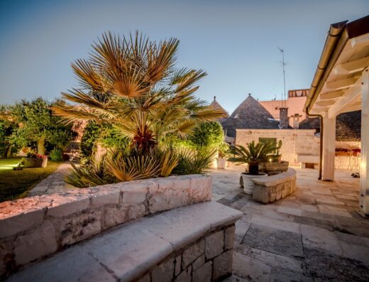 Astra B&B Alberobello: Veduta esterna serale di un trullo con giardino illuminato, palme e sedute in pietra, tipico alloggio turistico pugliese.
