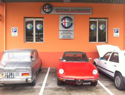 Autofficina Marconi Specializzata: Facciata arancione con insegne Alfa Romeo, parcheggio con Alfa Romeo d'epoca grigia, rossa spider e auto bianca con cofano aperto per riparazioni.