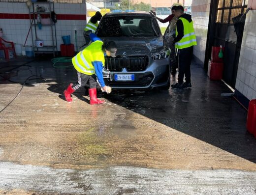 Autolavaggio a mano: Operatori lavano manualmente una BMW grigia in un autolavaggio, utilizzando spugne e acqua saponata per pulire la carrozzeria. Servizio professionale di pulizia auto.