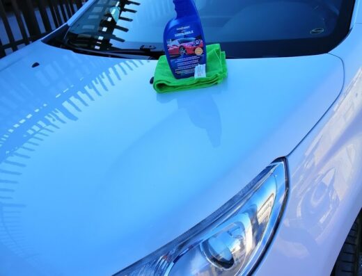 Autolavaggio a mano: Dettaglio del cofano di un'auto bianca con un flacone di detergente e un panno verde per la pulizia.