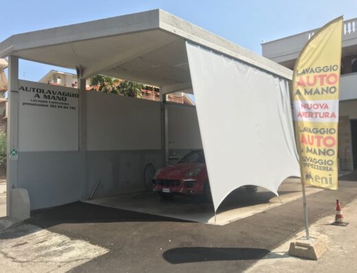 Autolavaggio a mano Eni: auto rossa in lavaggio, insegna 'Autolavaggio a mano' e bandiera pubblicitaria con scritta 'Lavaggio auto a mano'.