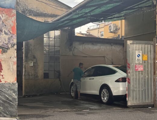 Autolavaggio a mano "Il Siluro Carrara": Un uomo lava a mano una Tesla bianca, servizio di autolavaggio artigianale in un ambiente esterno.