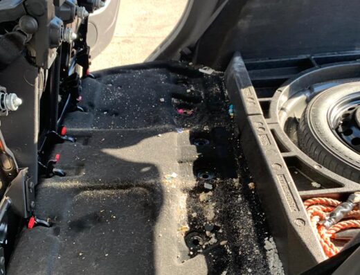 Interno sporco del bagagliaio di un'auto che necessita di autolavaggio a mano e pulizia a vapore, con sedili reclinati e ruota di scorta visibile.