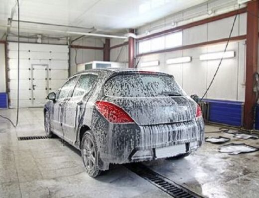 Un'auto grigia è ricoperta di schiuma di sapone in un autolavaggio Almax, con scarichi a griglia visibili nel pavimento.