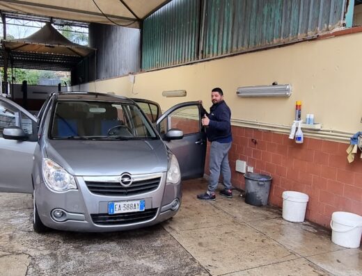 Autolavaggio Car Wash: Un uomo lava la sua auto grigia, una berlina, in un autolavaggio all'aperto. Secchi e prodotti per la pulizia sono visibili. Servizio di lavaggio auto self-service.