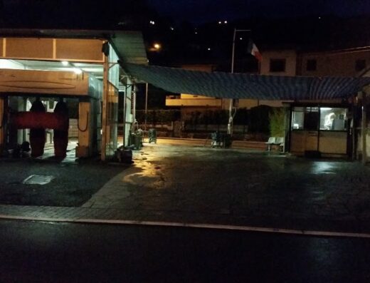 Autolavaggio CM: Foto notturna dell'autolavaggio con struttura per il lavaggio auto, tettoia, cabina di servizio e bandiera italiana sullo sfondo.