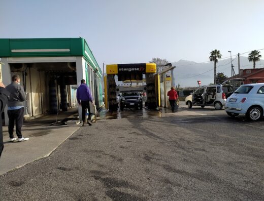 Autolavaggio Del Freo: Vista esterna dell'autolavaggio con auto in fase di lavaggio, persone in attesa e veicoli parcheggiati.