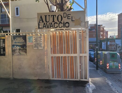 Autolavaggio Del Sole Busiello: Entrance to the car wash service with signage, under a corrugated metal awning, featuring a metal gate with orange accents on a sunny day.