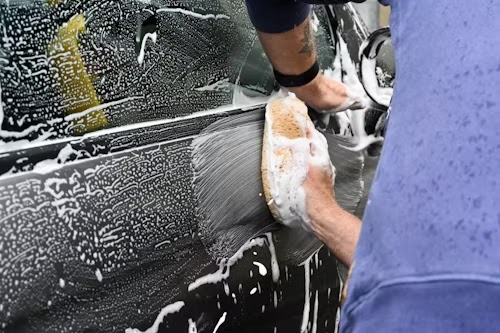 Un addetto lava a mano una macchina nera all'Autolavaggio di Poma Marcello & C. sas, usando una spugna e sapone per pulire la carrozzeria.