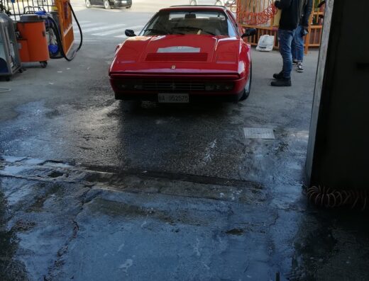 Un'auto sportiva rossa, probabilmente una Ferrari, in un autolavaggio DS2, con persone che la puliscono e l'ambiente circostante bagnato.