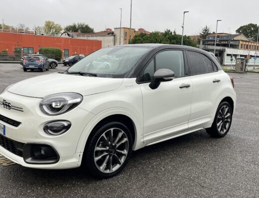 Fiat 500X bianca parcheggiata in un'area di servizio, con cielo nuvoloso. Accessori auto e dettaglio degli pneumatici visibili.