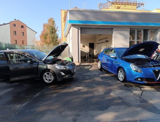 Due automobili con il cofano aperto all'Autolavaggio Fabio sas Eni: una Ford Focus grigia e una Alfa Romeo Giulietta blu, in attesa di manutenzione.