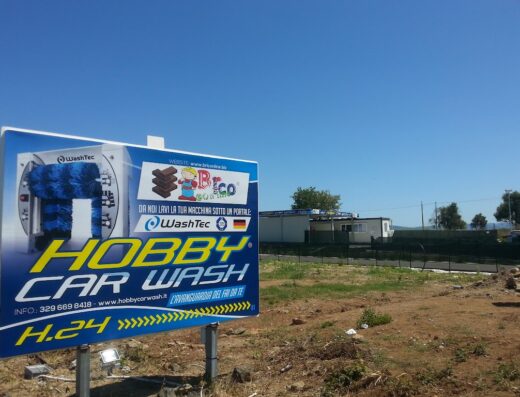 Cartello pubblicitario di Autolavaggio Hobby Car Wash, con l'immagine di un impianto di lavaggio auto WashTec, logo Hobby Car Wash e informazioni di contatto. Servizio di lavaggio auto self-service.