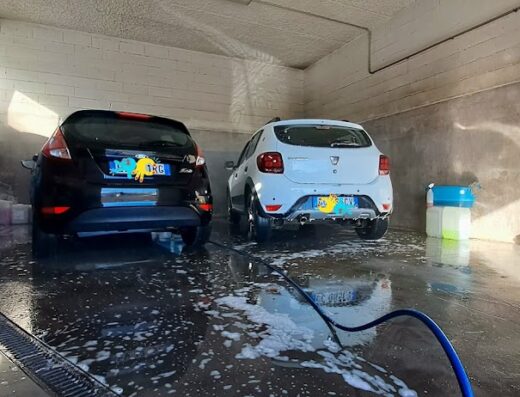 Due auto, una Ford Fiesta nera e una Dacia Sandero bianca, in un autolavaggio, con schiuma e acqua sul pavimento.