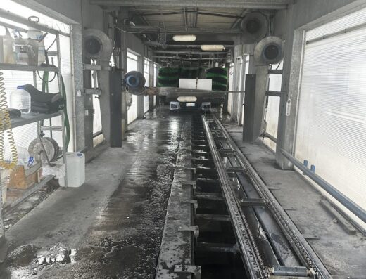 Interno dell'autolavaggio Nouvelle Olimpia, con attrezzature per il lavaggio moto e veicoli, binari a terra e spazzole rotanti visibili.