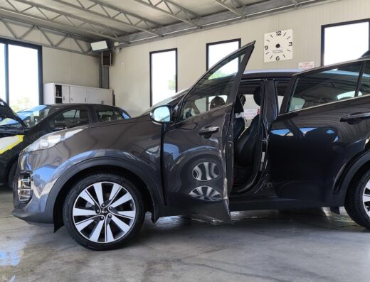 Auto grigia in autolavaggio con portiera aperta, riflessi sul pavimento bagnato, dettaglio cerchioni e interni, orologio a muro.