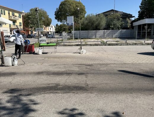 Autolavaggio Quadrifoglio: A man stands near a table with a sign that says "Aperto" at an outdoor location, possibly offering motorcycle cleaning services.