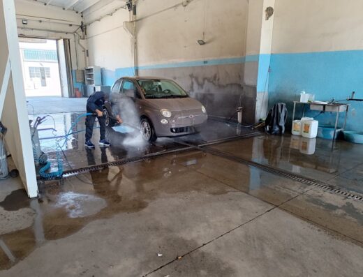 Autolavaggio rapido a mano: un addetto lava una Fiat 500 grigia con idropulitrice professionale in un autolavaggio.