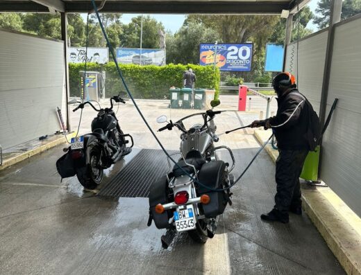 Autolavaggio Scapecchi: Due moto, una nera e una grigia, vengono lavate in un'area self-service. Un uomo pulisce la moto grigia con una lancia ad alta pressione. Accessori da viaggio visibili sulle moto.