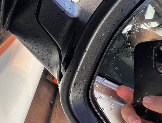 Dettaglio dello specchietto retrovisore di un'auto, bagnato con gocce d'acqua, durante un autolavaggio self-service. Si intravede anche una mano che impugna un telefono.