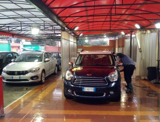 Autolavaggio Serena: Mini Cooper blu scuro sottoposta a lavaggio auto professionale, con operatore che la pulisce. Pavimento bagnato.