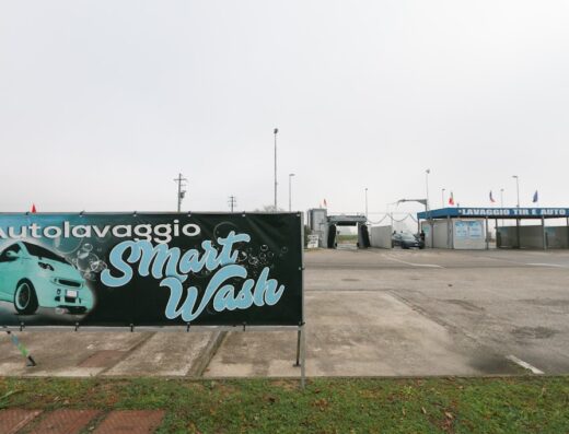 Insegna dell'autolavaggio Smart Wash con un'auto stilizzata e la scritta "Autolavaggio Smart Wash" in evidenza, con sullo sfondo la struttura self-service per il lavaggio di auto e furgoni.