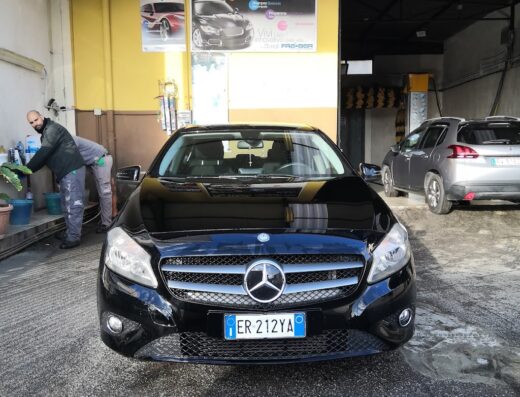 Primo piano di una Mercedes nera all'Autolavaggio Stracuzzi, con operai che lavorano sullo sfondo. Servizi professionali per la cura dell'auto.