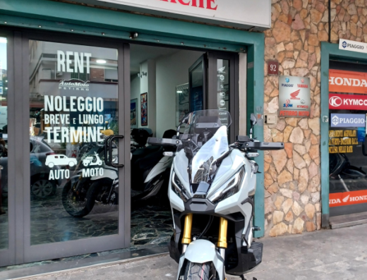 Automoto Petirro: scooter Honda ADV350 parcheggiato di fronte al negozio con cartello "Noleggio Auto e Moto breve e lungo termine". Servizi di vendita e noleggio moto.