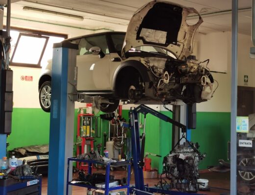 Un'auto bianca sollevata su un ponte in un'officina di autoservizio, con il cofano aperto e il motore parzialmente smontato, pneumatici impilati in primo piano.